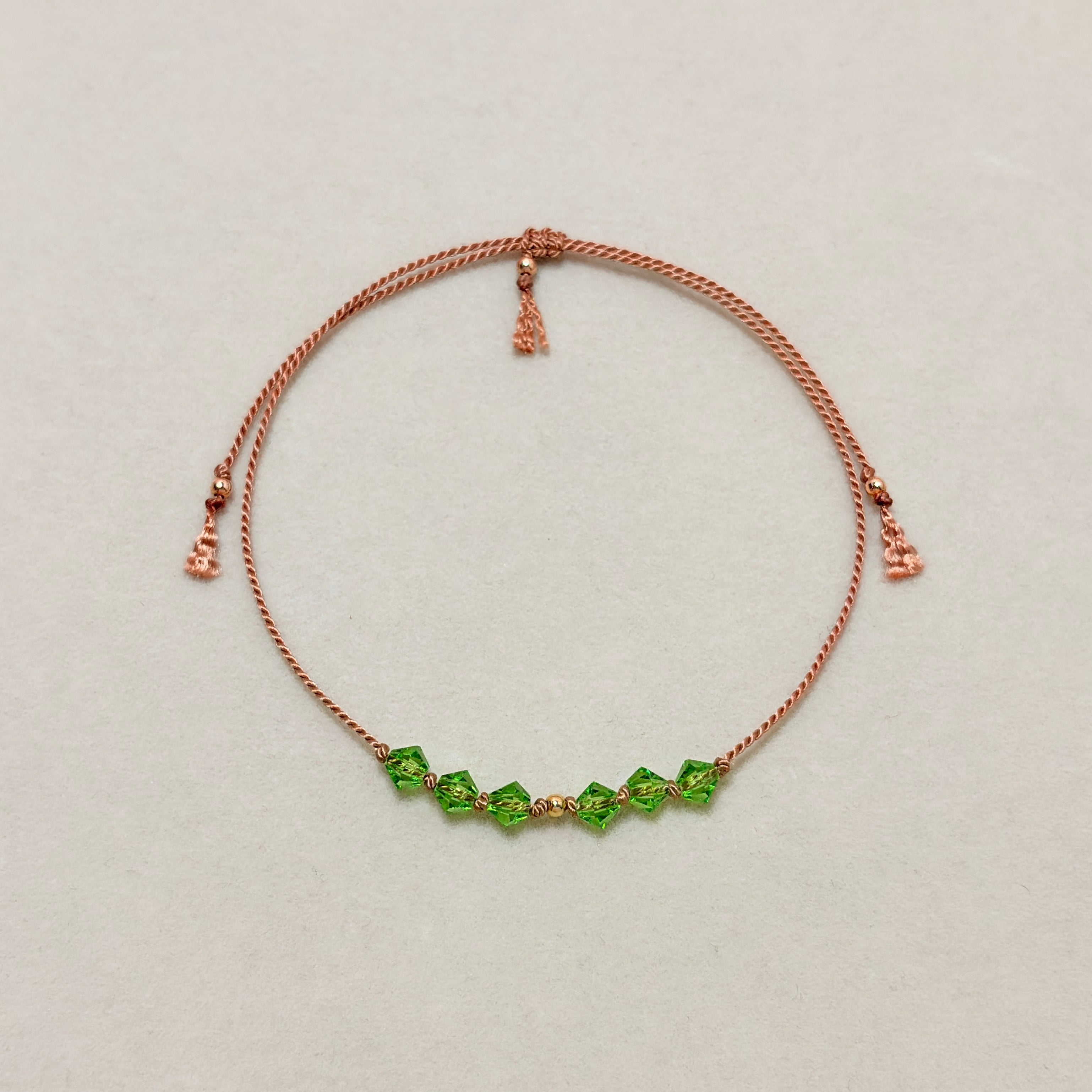 Renewal adjustable bracelet with green Peridot crystals, beige silk cord, and gold-filled details on a soft ivory velvet background.
